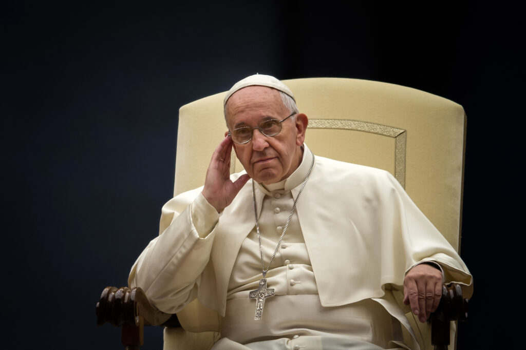 The Pope sits on a papal chair dressed in white and with is head resting on his right hand.