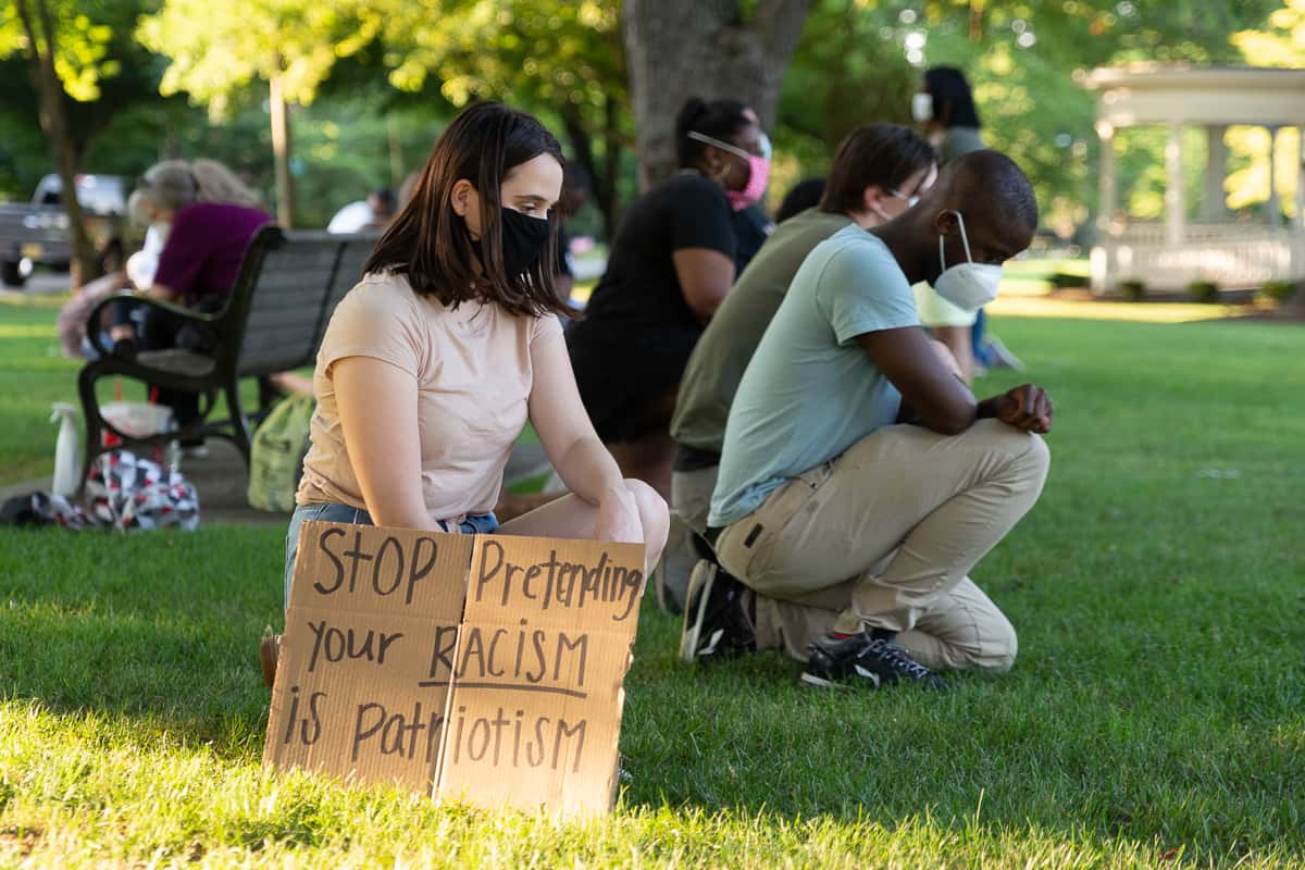 A young woman kneels at a protest behind a cardboard sign on which is written "Stop pretending that your racism is patriotism."