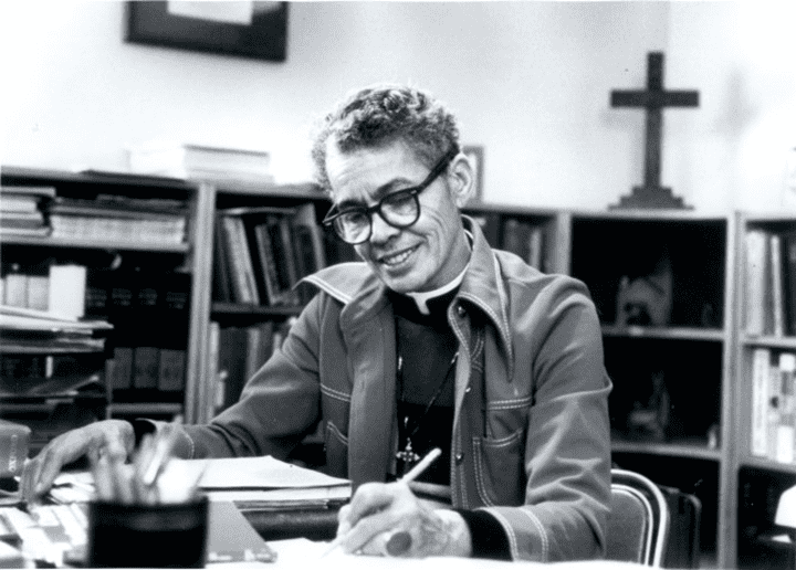 Episcopal Priest and civil right leader Pauli Murray sits at a desk in an office with a pen in hand and a smile on her face in this black and white photo.