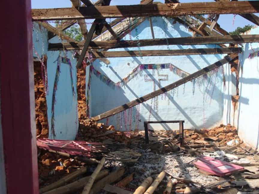 Photo shows what was once an Orissa church with the roof missing and daylight streaming through timbers. The outline of a cross is shown on a blue wall.