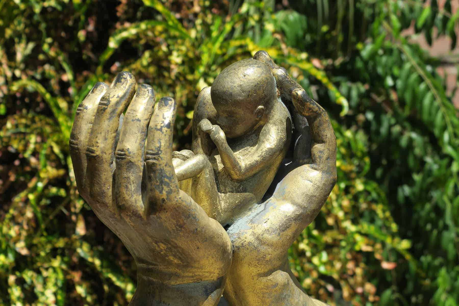 Hands of a sculpture are shown wrist together and holding a tiny baby.