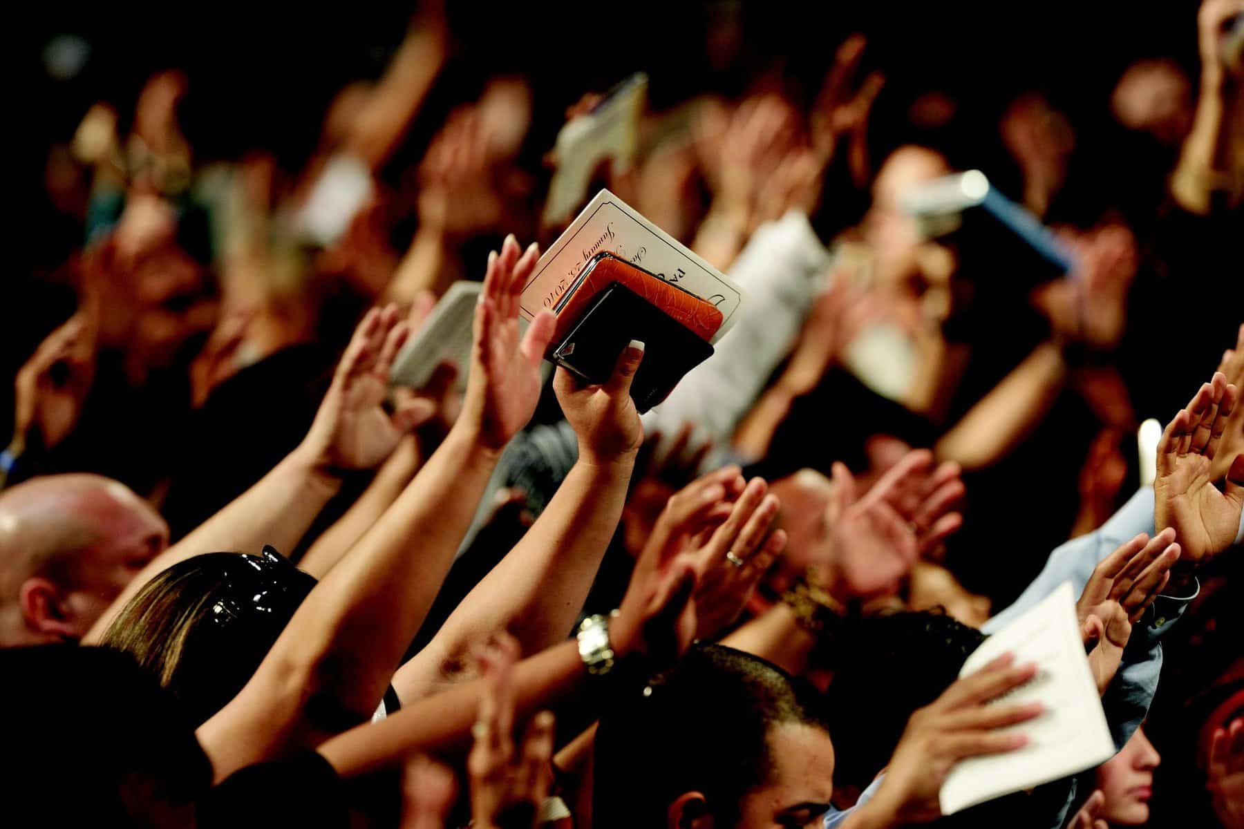 Hands a bibles are raised up in a large meeting space, perhaps a church.