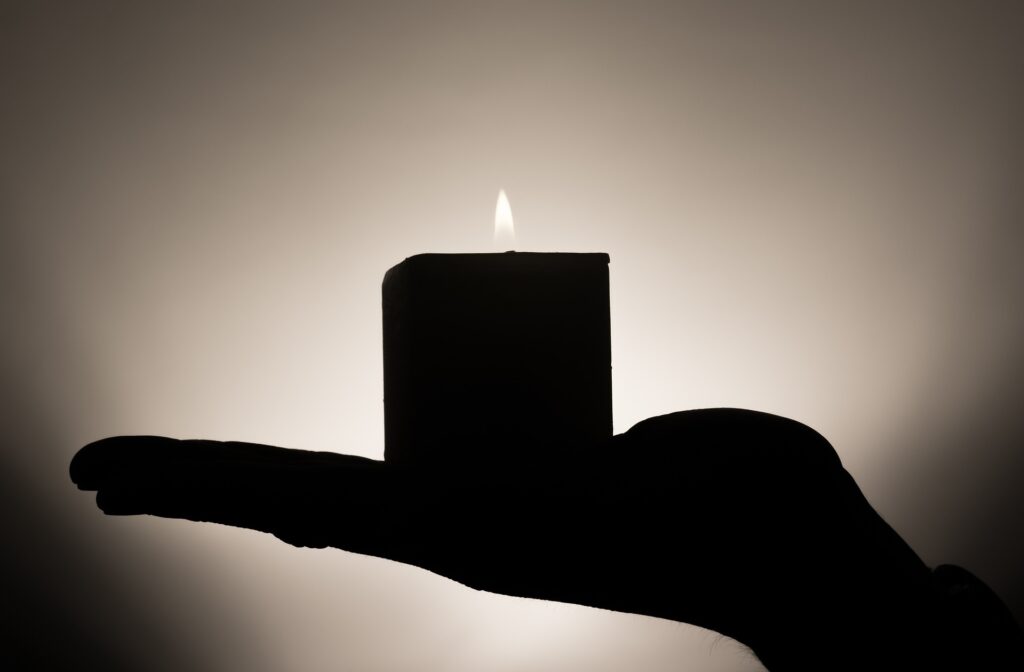 A hand is shown in silhouette holding a short, lit, pillar candle.