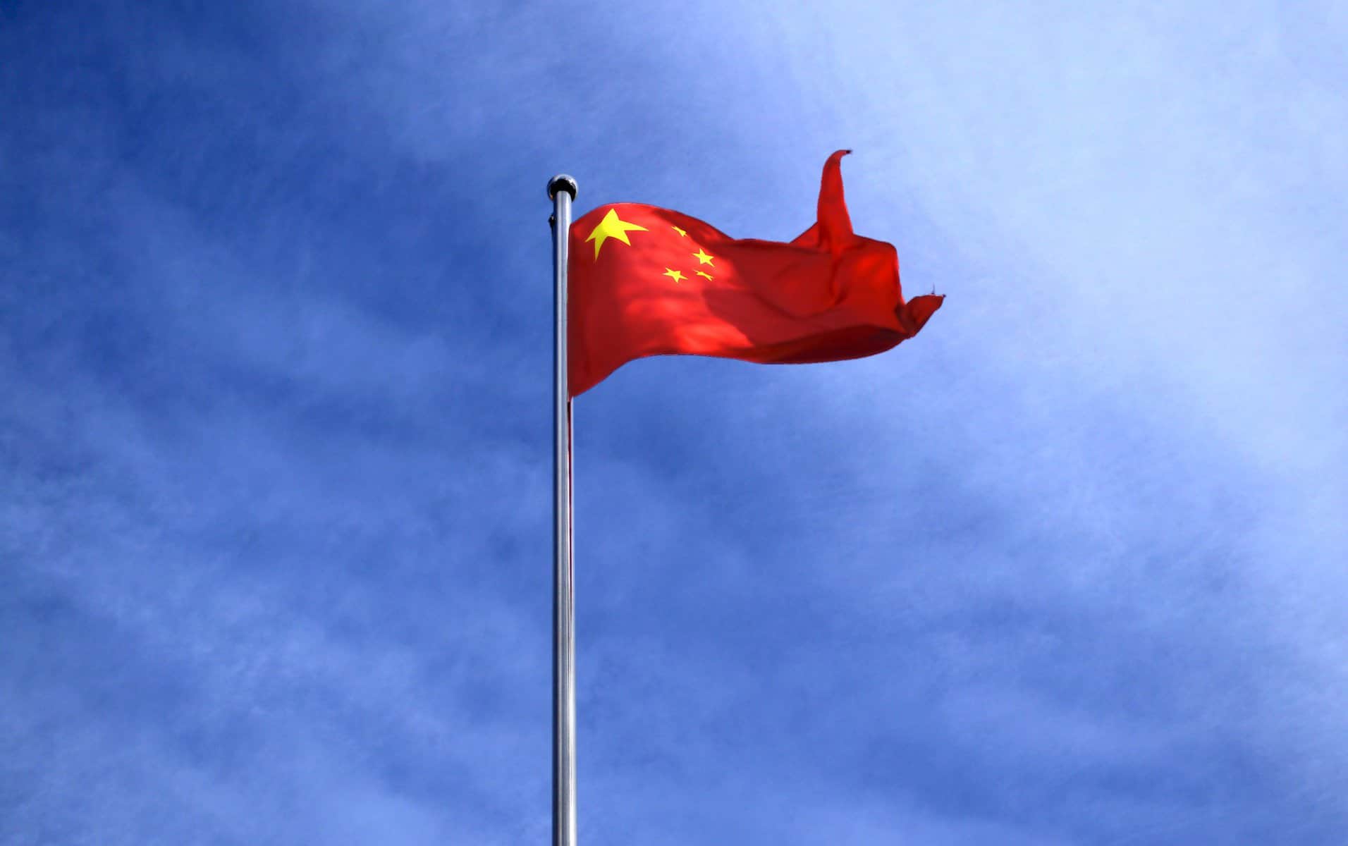 The flag of China waves in the breeze atop a flagpole against a blue sky.