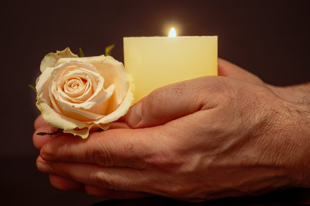 A Caucasian man's hands are shown holding a lit candle in a votive and and white rose.