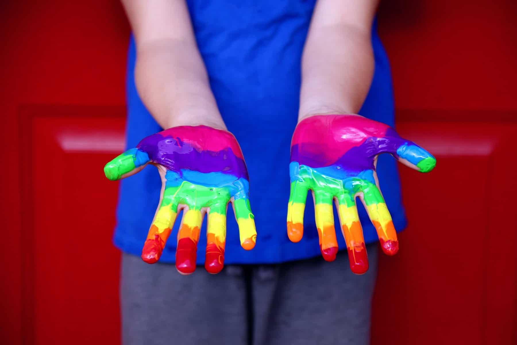 A child stands in front of a red garage door in a blue t-shirt with palms up and painted in rainbow colors.