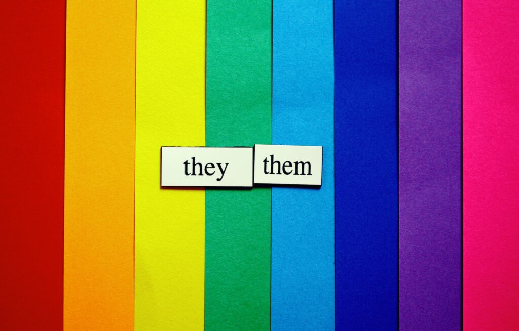 Paper rainbow stripes are overlaid with two pieces of paper, one saying they, the other them.