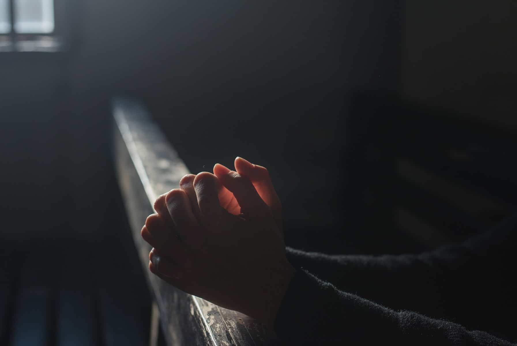 Hands are clasped in prayer at an altar rail with sunlight backlighting them through a window.