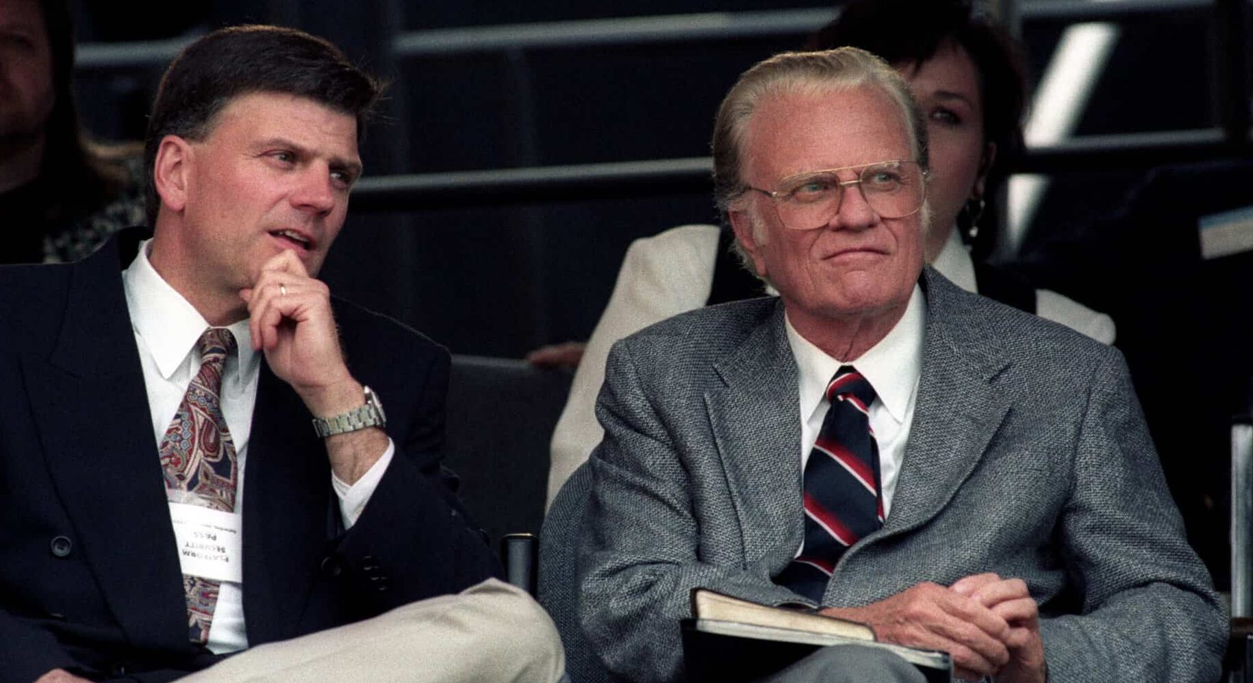 Billy Graham is shown seated in an auditorium type setting and wearing a grey suit.