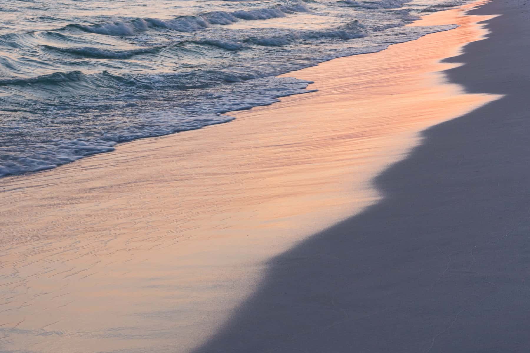 Ocean waves are shown sliding back into off of sand with the newly soaked sand reflecting the pink sunset light.