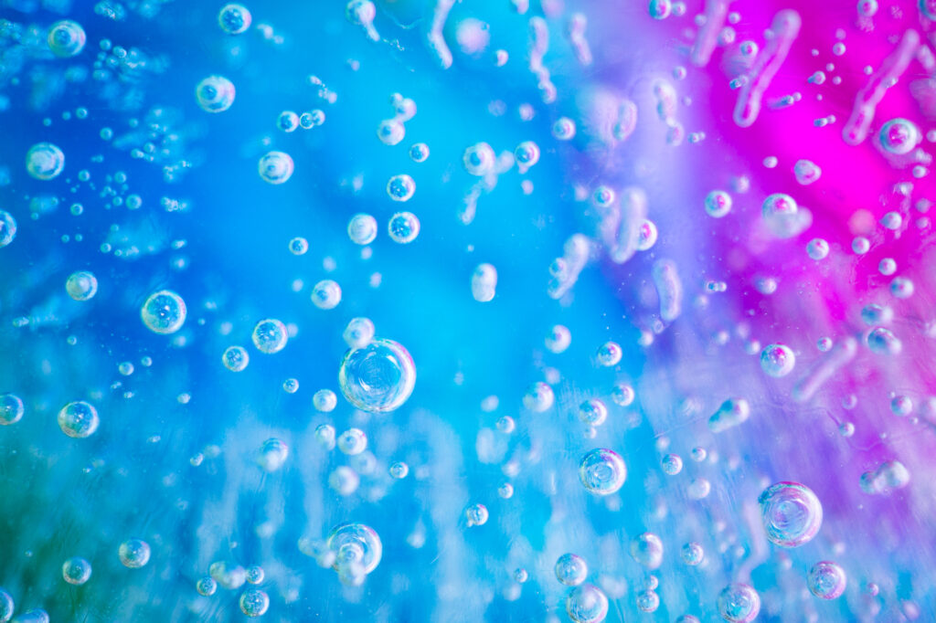 This macro photo shows ice water with many suspended frozen bubbles against a backdrop of mostly blue with some pink and green.