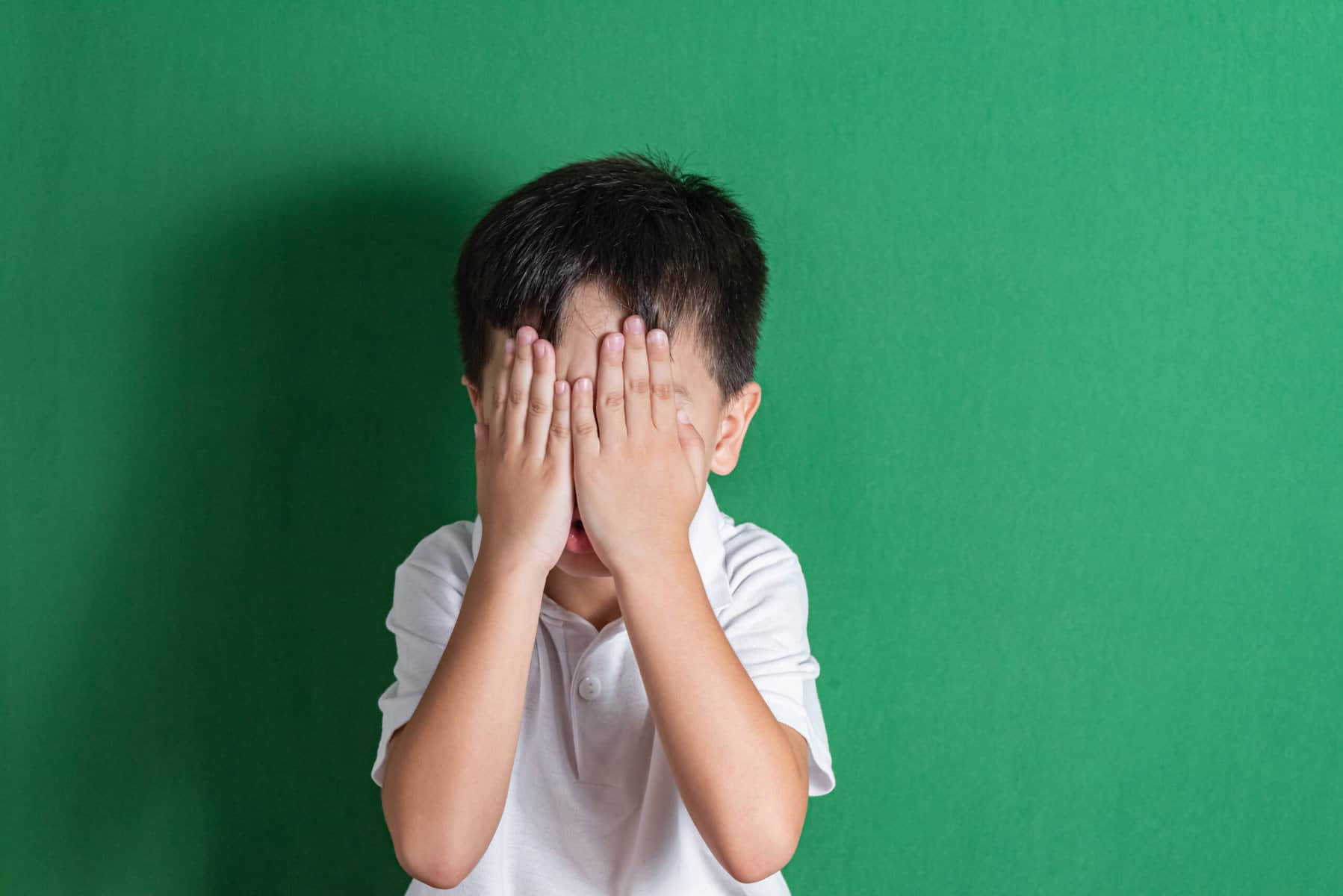 A child is shown in a white polo shirt in front of a vibrant green wall with his hands totally covering his face.