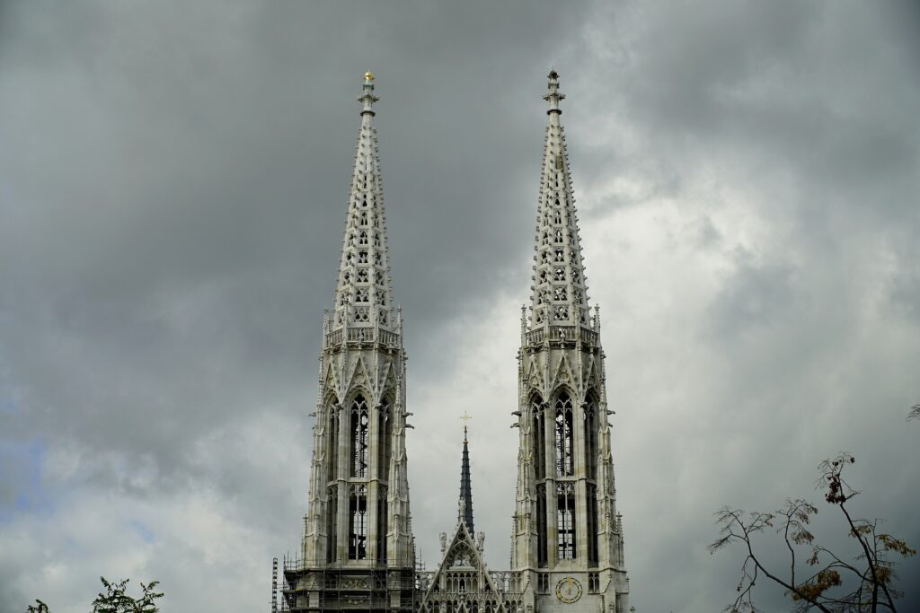 Two tall, thin white stone spires of a church are shown against a cloudy sky.
