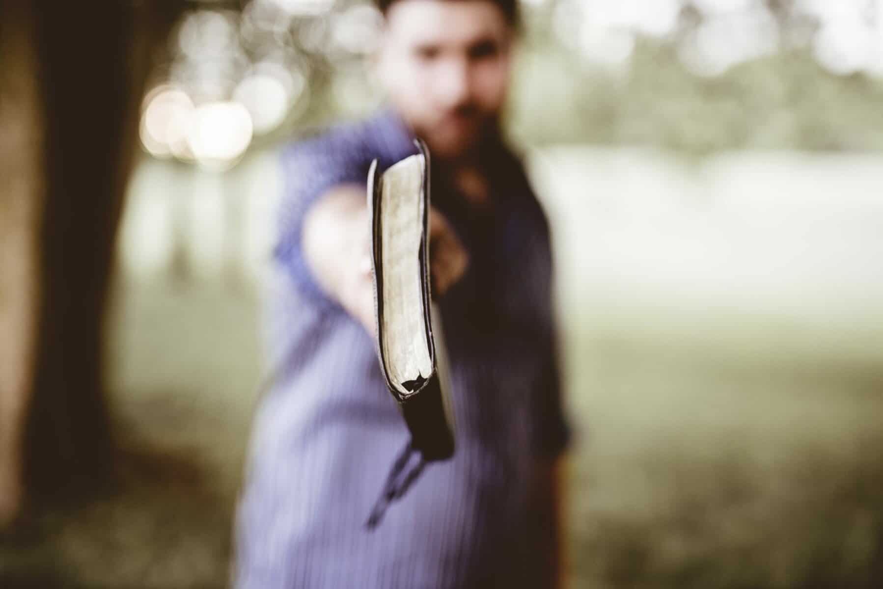 An out of focus many holds an in focus bible in front of himself at arm's length.
