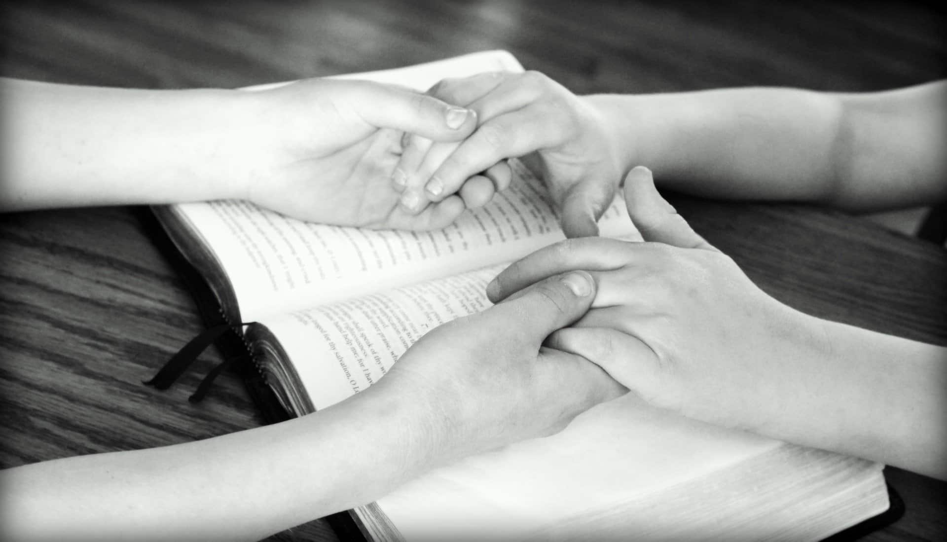 Black and white image shows two people's hands a forearms. The people are holding hands resting on a bible that is open on a table top.