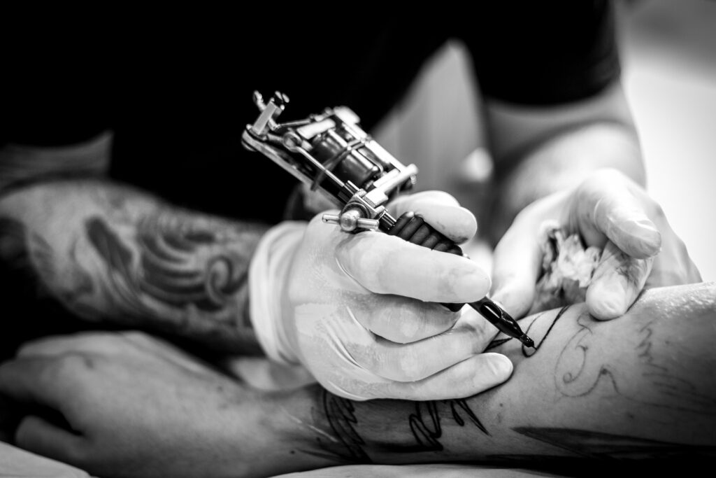 This black and white image shows the hands of a tattoo artist holding a tattoo gun and applying a tattoo to a customer's forearm.