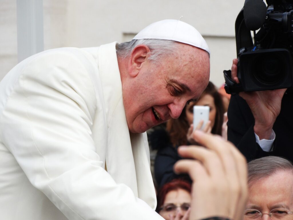 Pope Francis is shown bending over and talking to someone in a crowd.