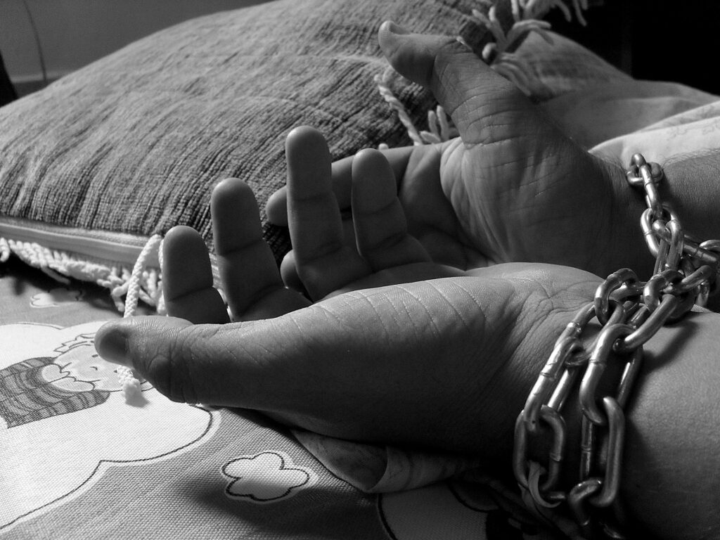 This black and white image shows a person's hands palm up, wrists bound in chains atop a bed with a small pillow.