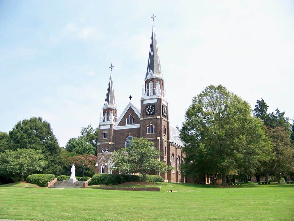 A red stone church with white stone accents and steep, pointed spires with green grass shown in the foreground.