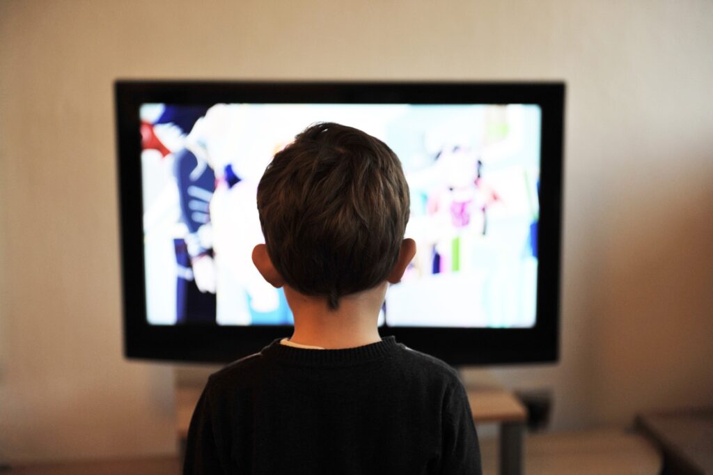 The back of a young boy's head is shown as he stands looking at an out-of-focus TV.