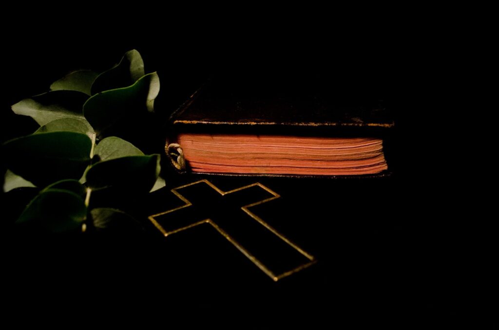 A black background shows a closed black bible with red page edges, a metal cross, and some plant leaves.