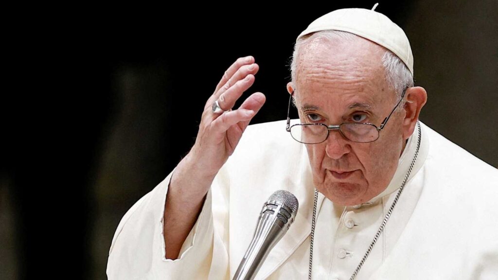 The Pope is seen in his papal robe and cap speaking at an event with his right hand gesturing upwards.