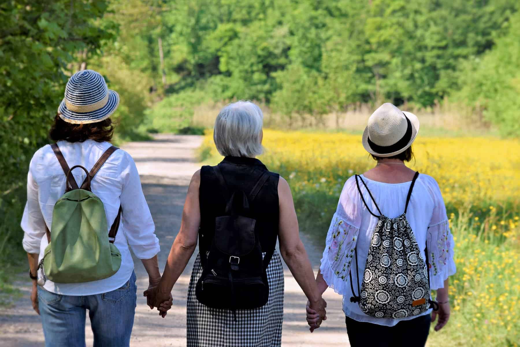 Three older women are shown wearing backpack purses and holding hands as they walk down a nature path.