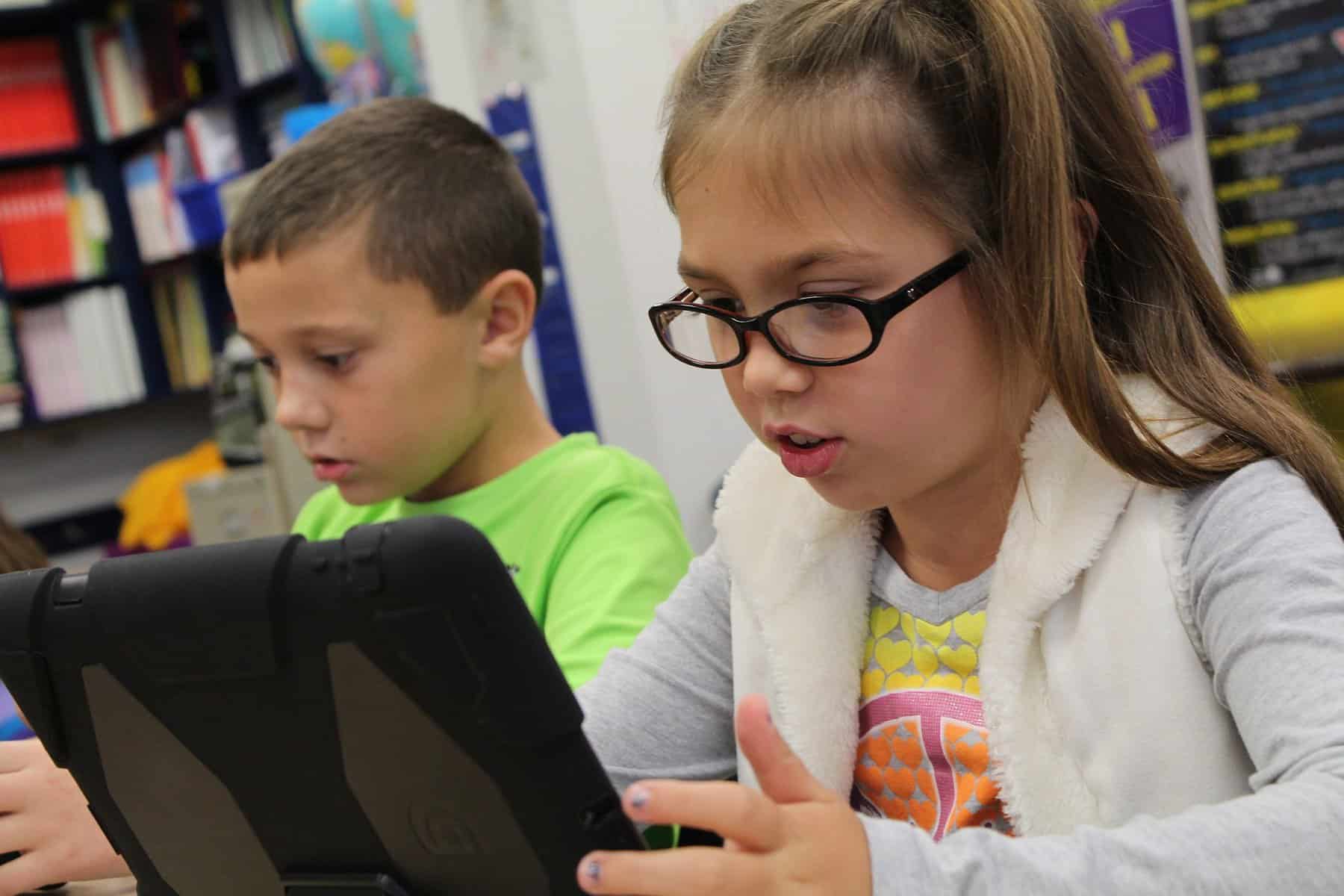 A young girl in glasses is seen looking at a tablet device in a classroom setting.
