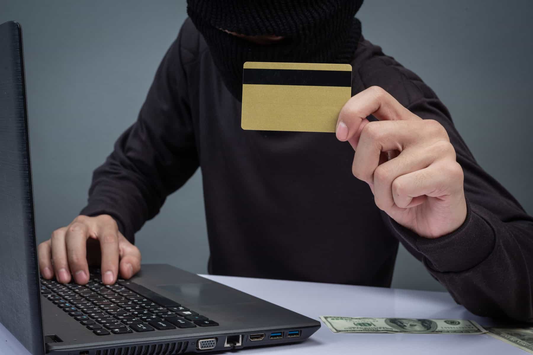 A person disguised in black clothing and a mask is seen as a computer with paper money at their elbow and holding a credit card.