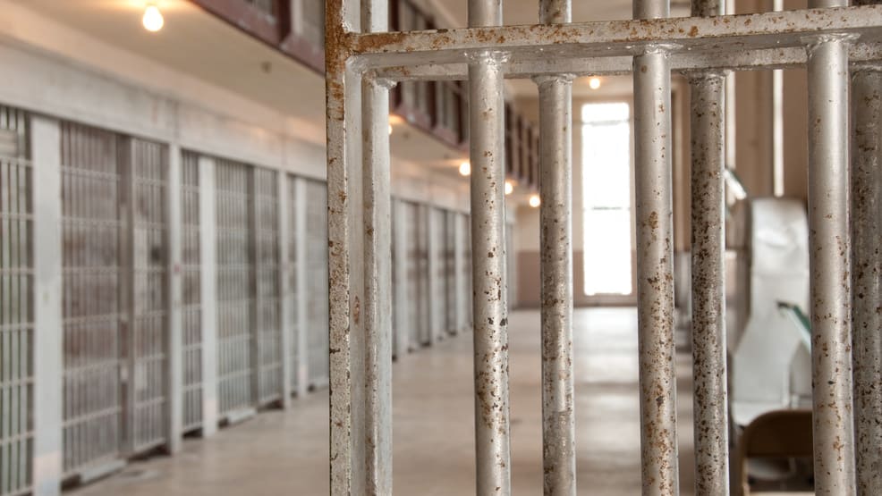 The inside of a prison is seen with cells at a distance with some bars close up.