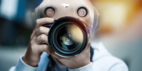 A camera and lens is held up and obscuring the face of a person with an apparent mask on.