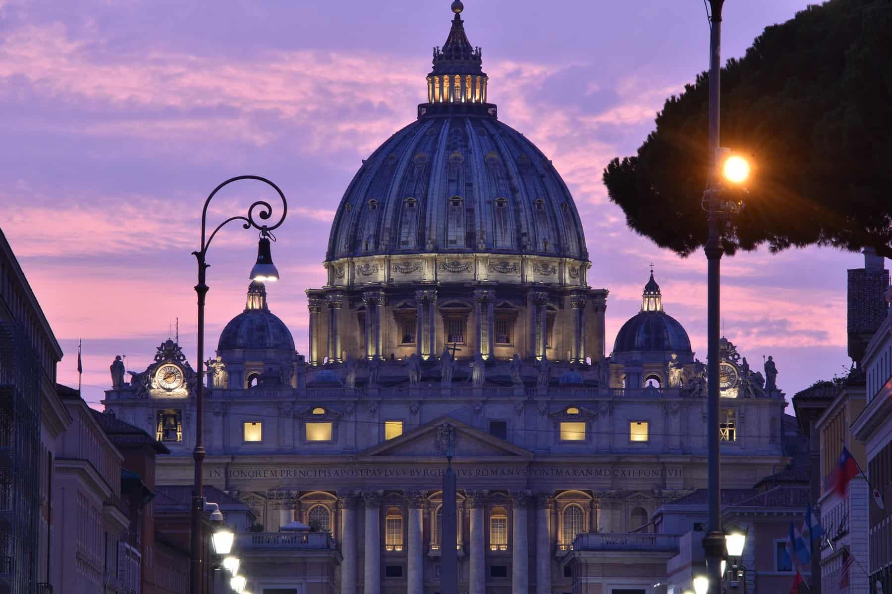 A lit dome in the Vatican is shown at sunset with pink and purple clouds behind it.