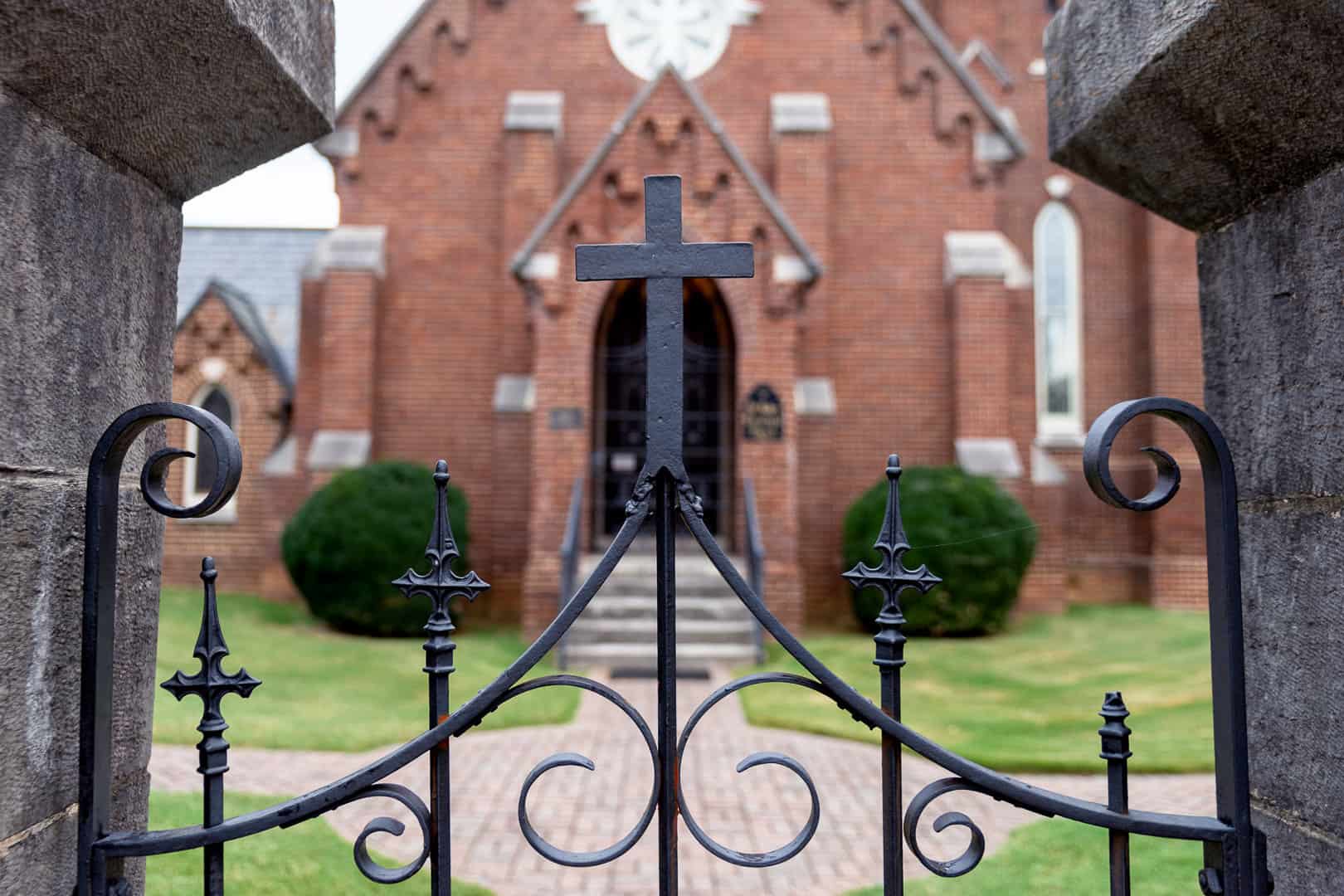 The chapel entrance with massive wooden doors and red brick and white stone entrance of St. Luke's Episcopal Church in Cleveland, TN is seen from the sidewalk and through the historic wrought iron sidewalk gate. The iron cross adorong the top of the gate is in focus and centered in the frame.