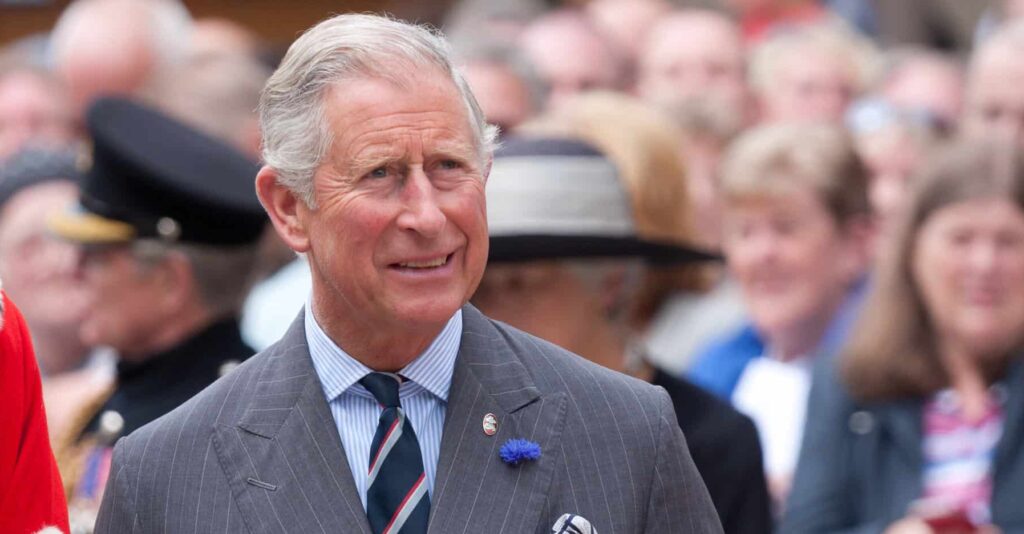 King Charles III is shown in gray suit and from the shoulders up with a crowd behind him.