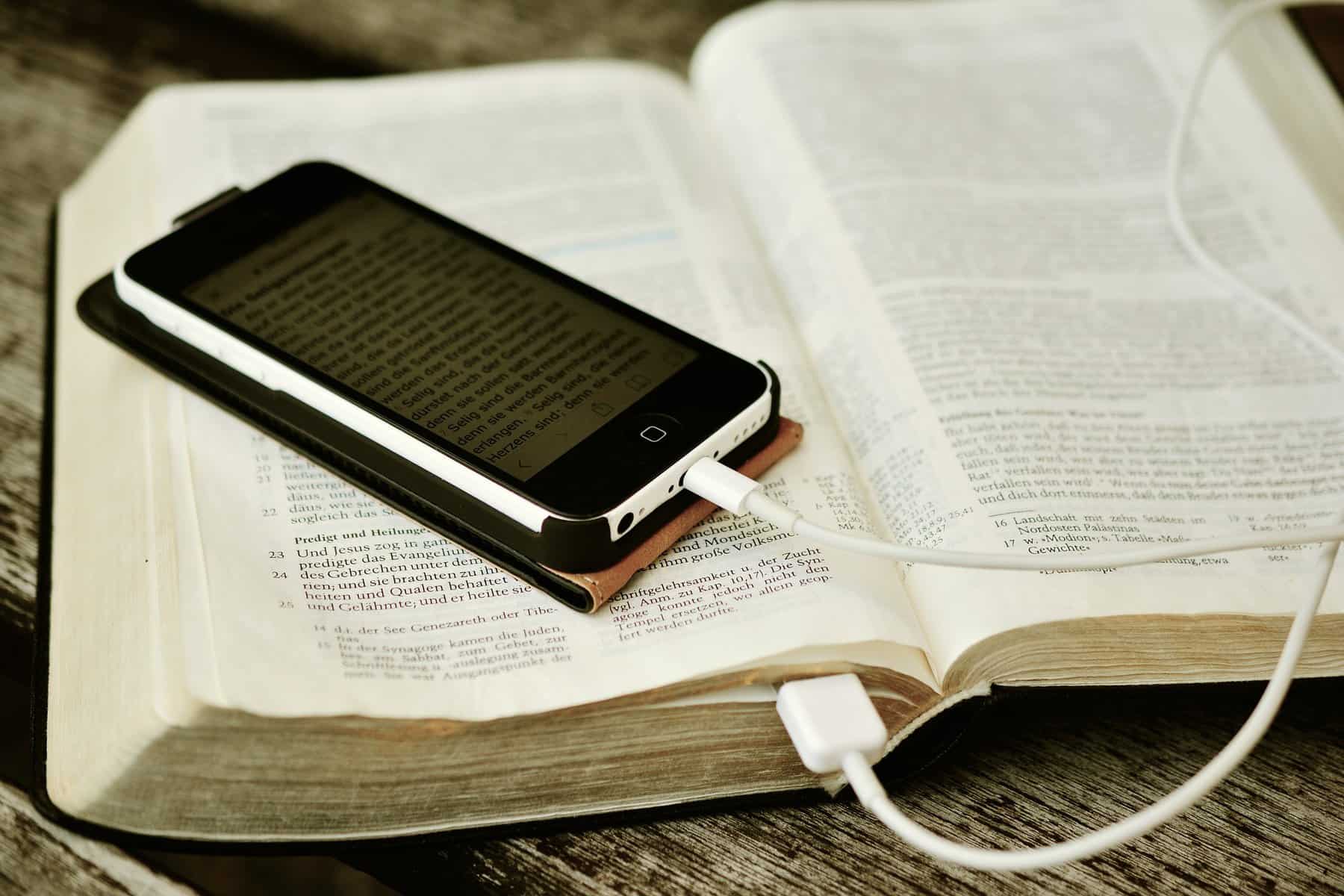 A smart phone is seen sitting on a bible with its cord appearing to be plugged into the open bible.