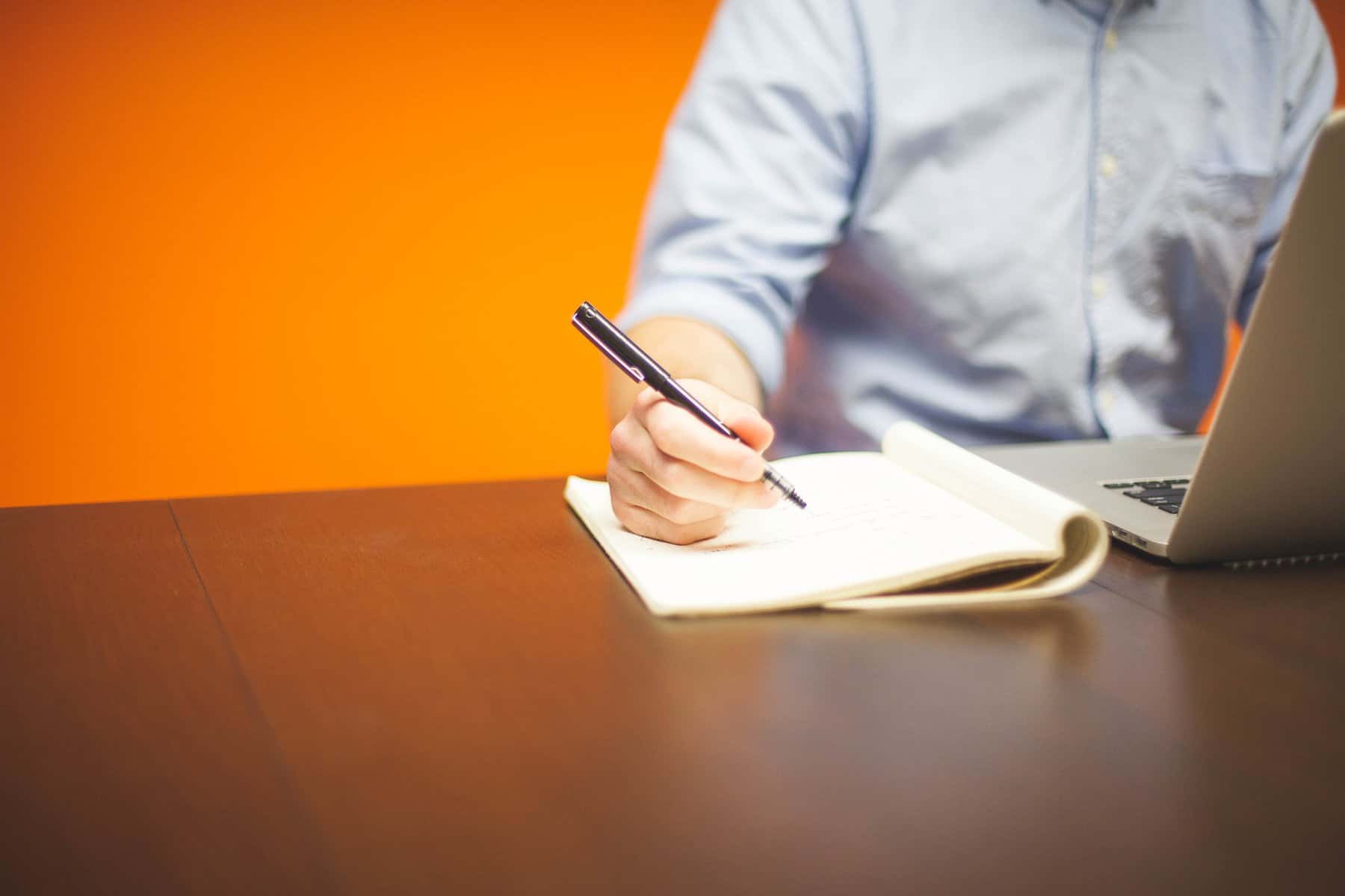 The torso of a man is shown sitting at a computer and writing on a pad of paper against an orange background.