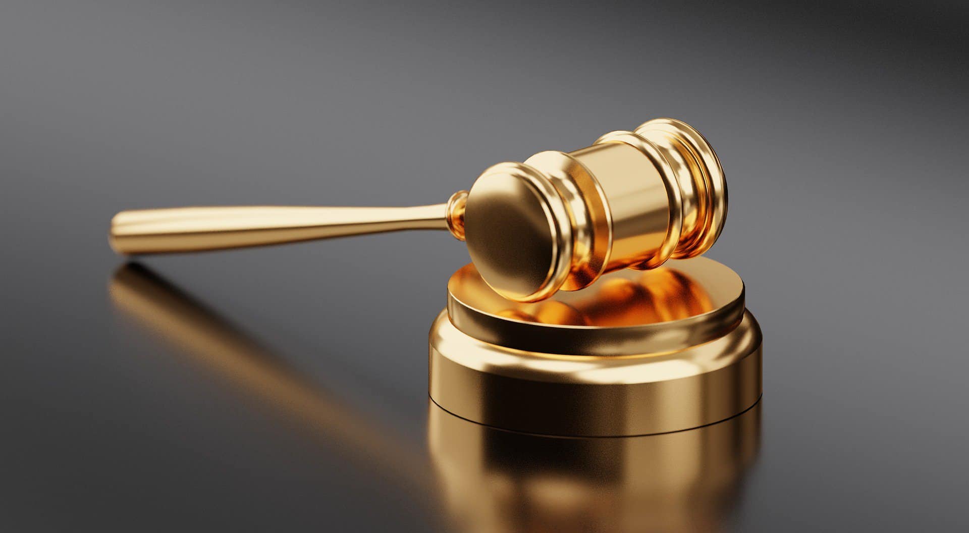 The head of a golden gavel sits on it's base on a dark reflective surface.