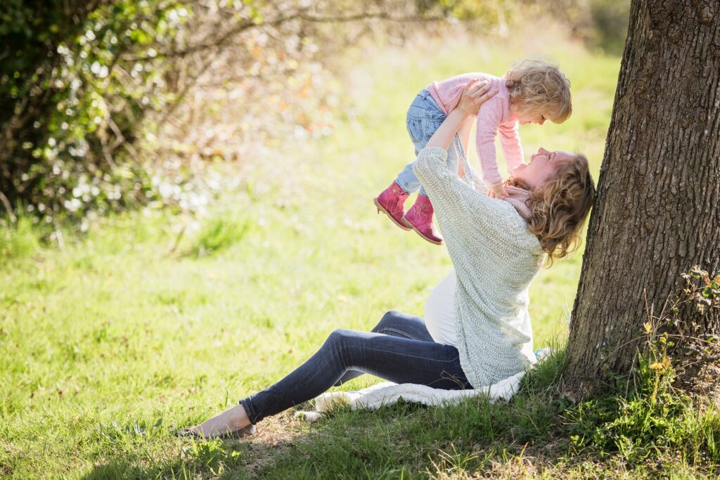 A woman is shown sitting against a tree and holding a toddler up in the air and both are smiling.
