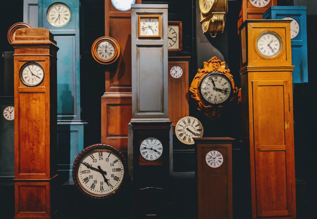 Grandfather clocks and mantle clocks of various colors and sizes are arranged all together in a darker room.