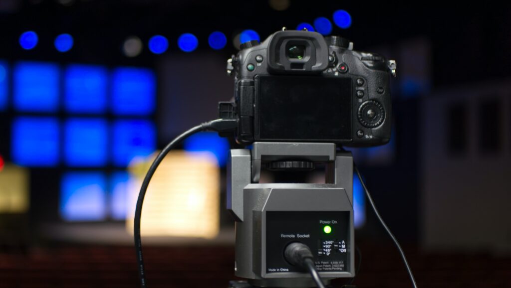 A camera for broadcasting is shown from the back facing a bank of blue screens.