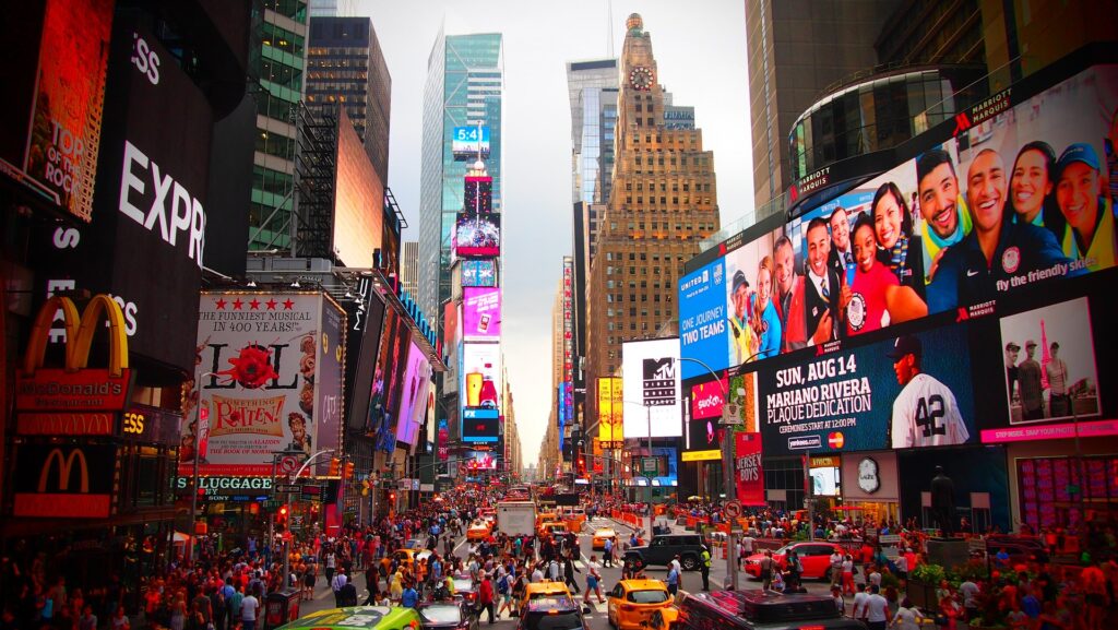 Times Square is seen from a couple stories high filled with people and jumbotrons.
