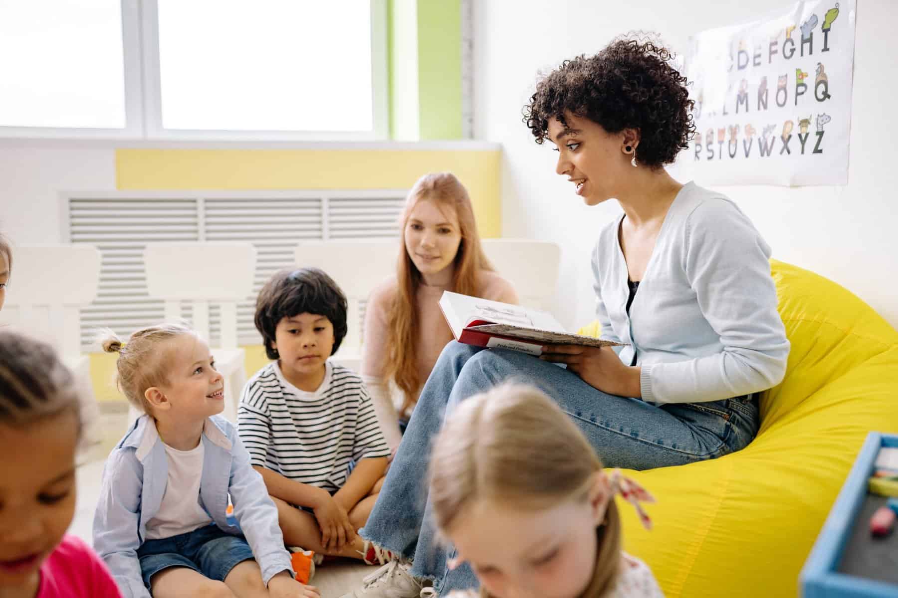 A woman sits in a yellow cushioned hair reading aloud to a group of young children.