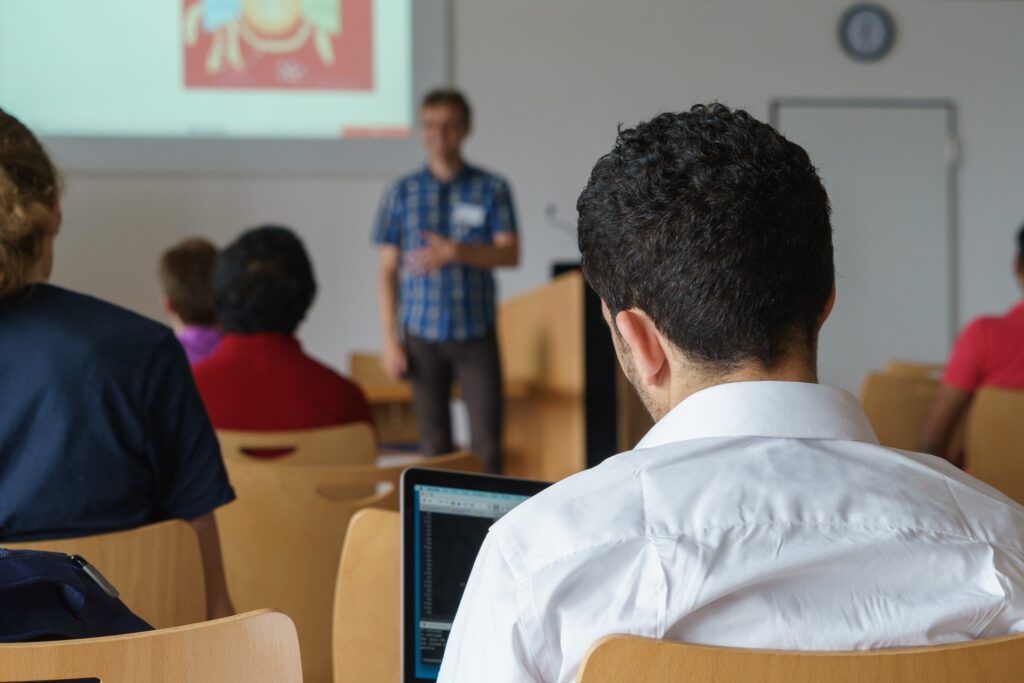 A student in a classroom is shown from the back with a teacher out of focus up front.