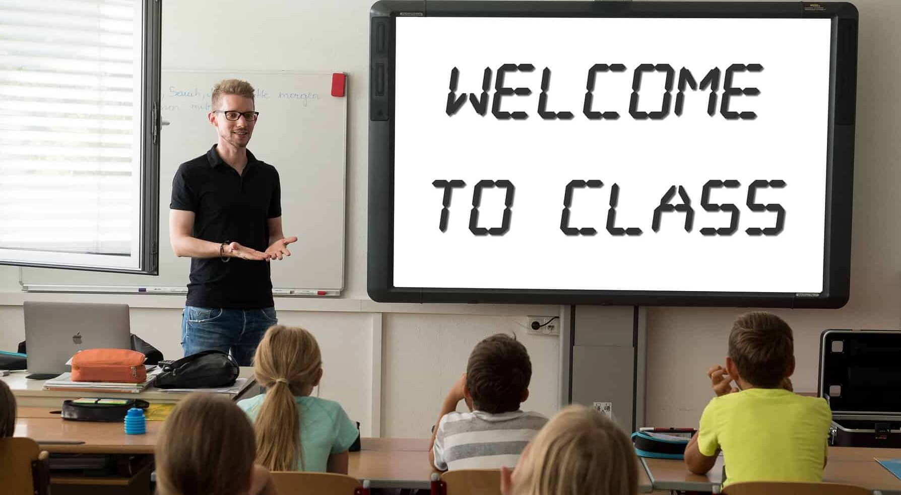 A male teacher stands at the front of a classroom of children with a sign saying "Welcome to Class".