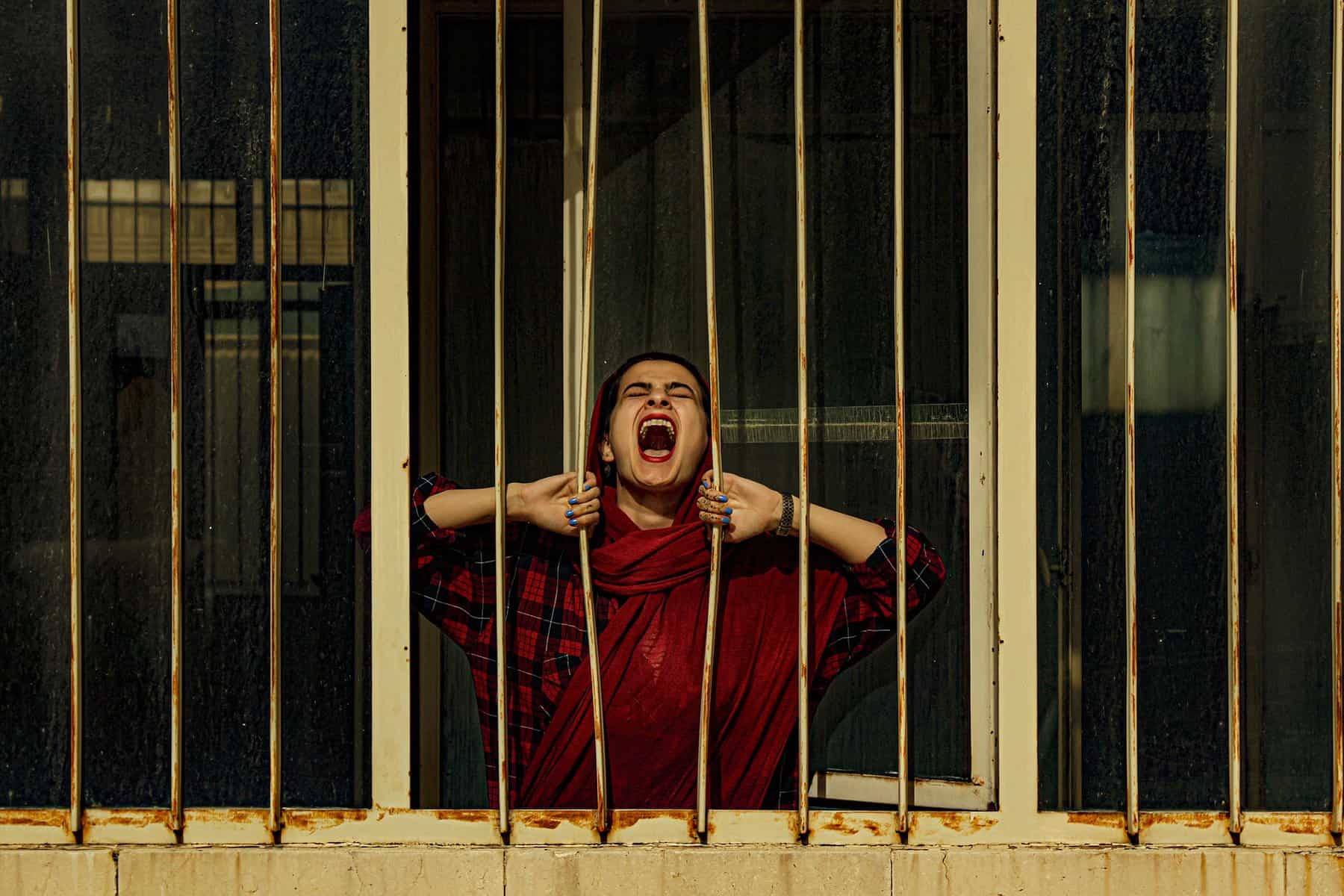 A woman in red is shown against bars with her hands trying to pull them apart while she yells.