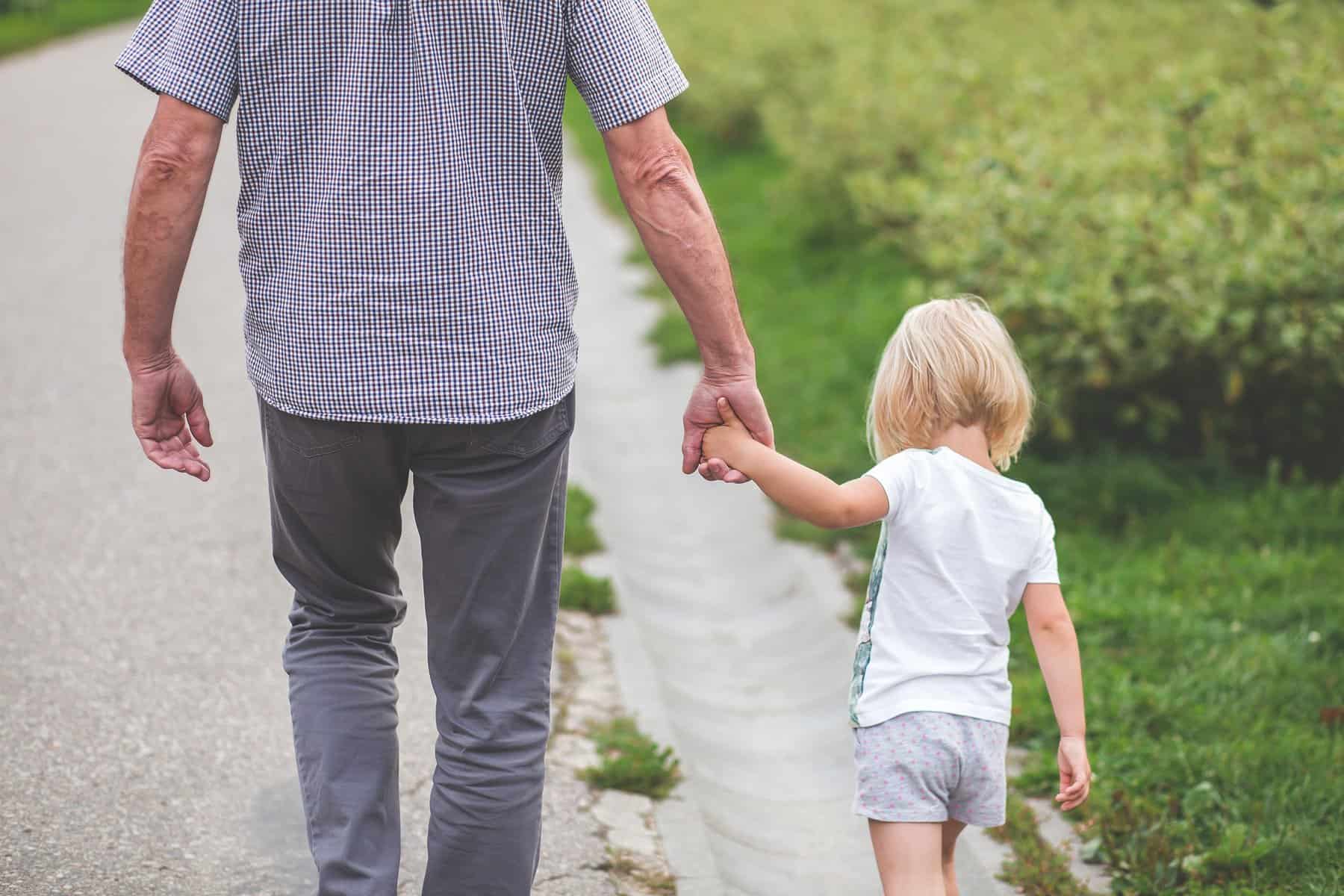 A man walks away from the camera holding the hand of a toddler as they walk side-by-side.