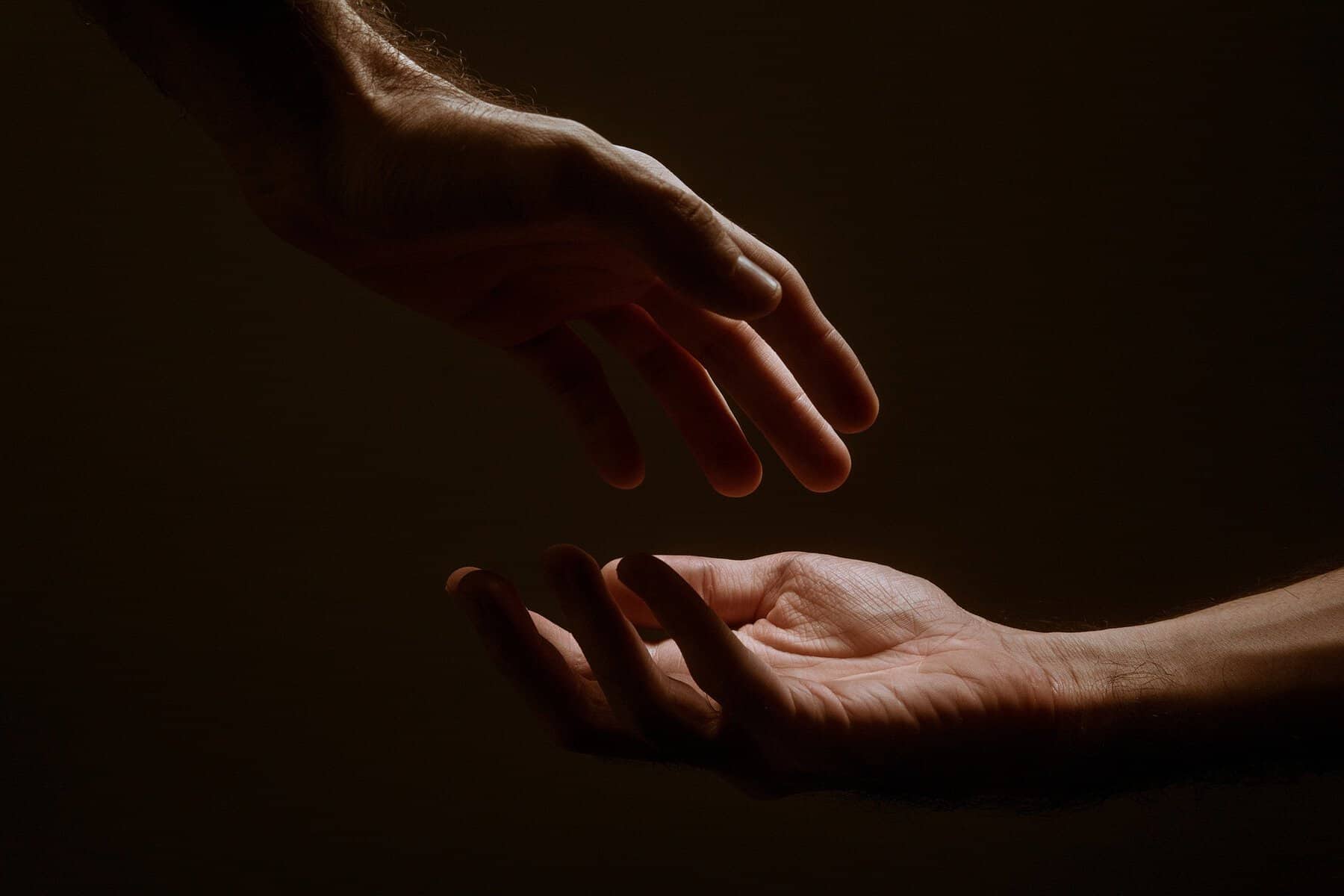 This moody image shows one hand extended out and above it another hand is reaching down toward it.