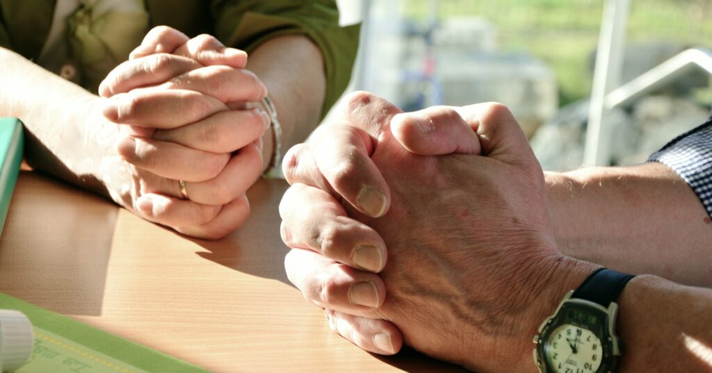The hands of two people are clasped and resting on a light colored wood table.