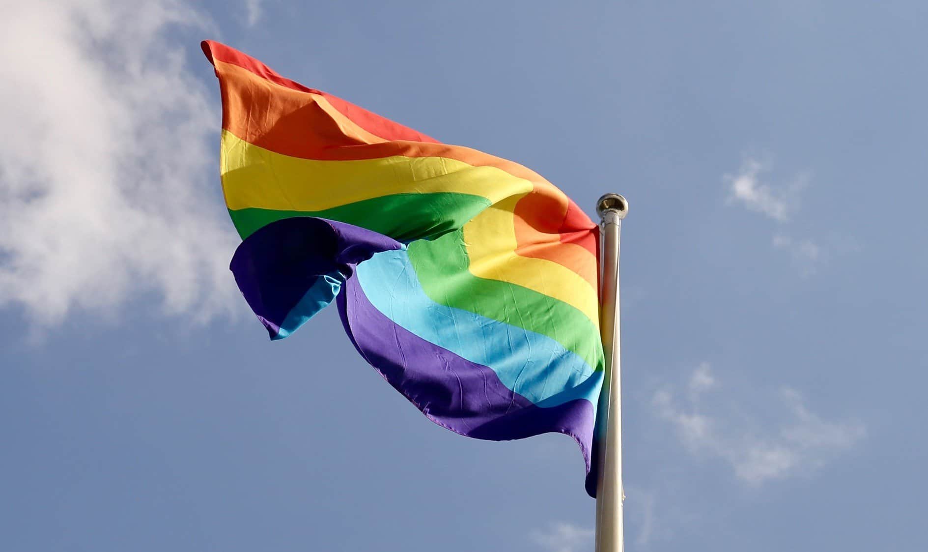 A rainbow Pride flag waves in the wind against a blue grey sky.