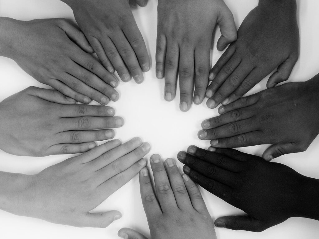 Many hands of multiple skin tones are placed flat in a circle on a white table in this black and white image.